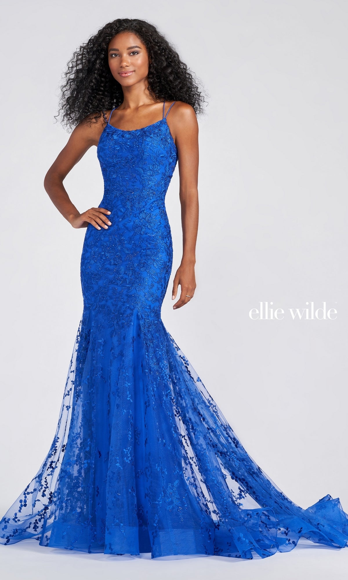A woman wears the Long Lace Mermaid Prom Dress with Strappy Back in royal blue, featuring thin straps, a fitted silhouette, and a sheer flared train. She stands against a light gray background with